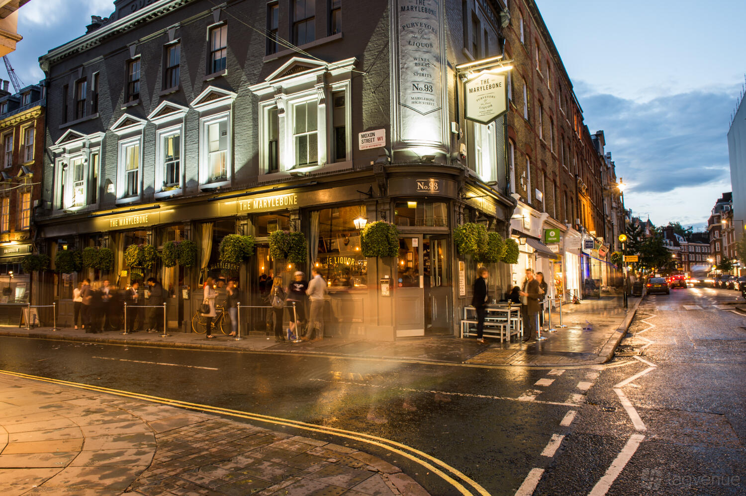 A corner pub with large windows, illuminated signage, and hanging greenery outside The Marylebone London.