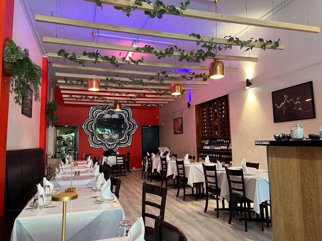 A restaurant with white linen tables, wooden chairs, hanging plants, and a wine rack at AAGAMAN – CBD.