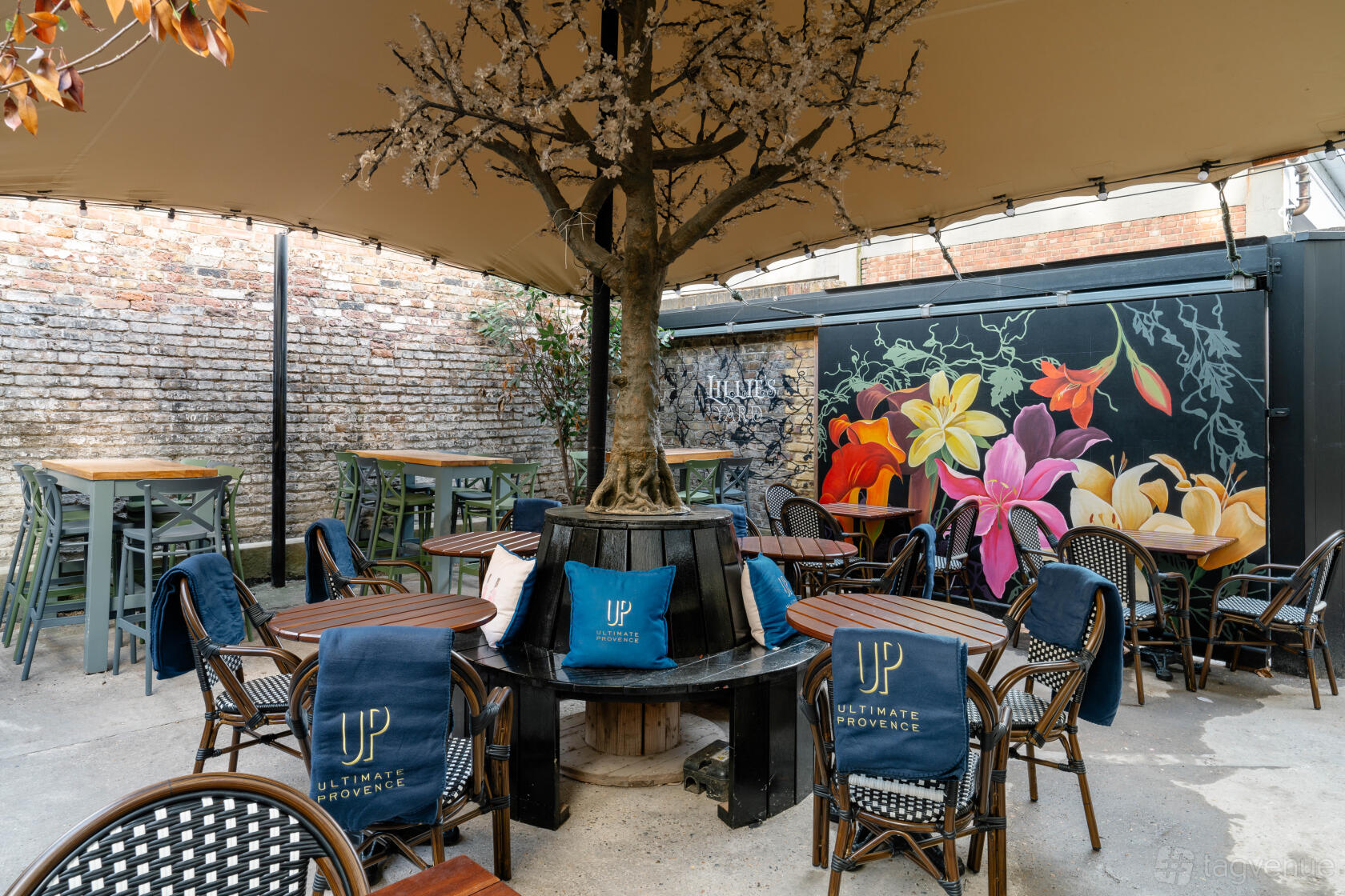 An open-air pub event space with a central tree, floral mural, and rattan chairs at The Lillie Langtry.