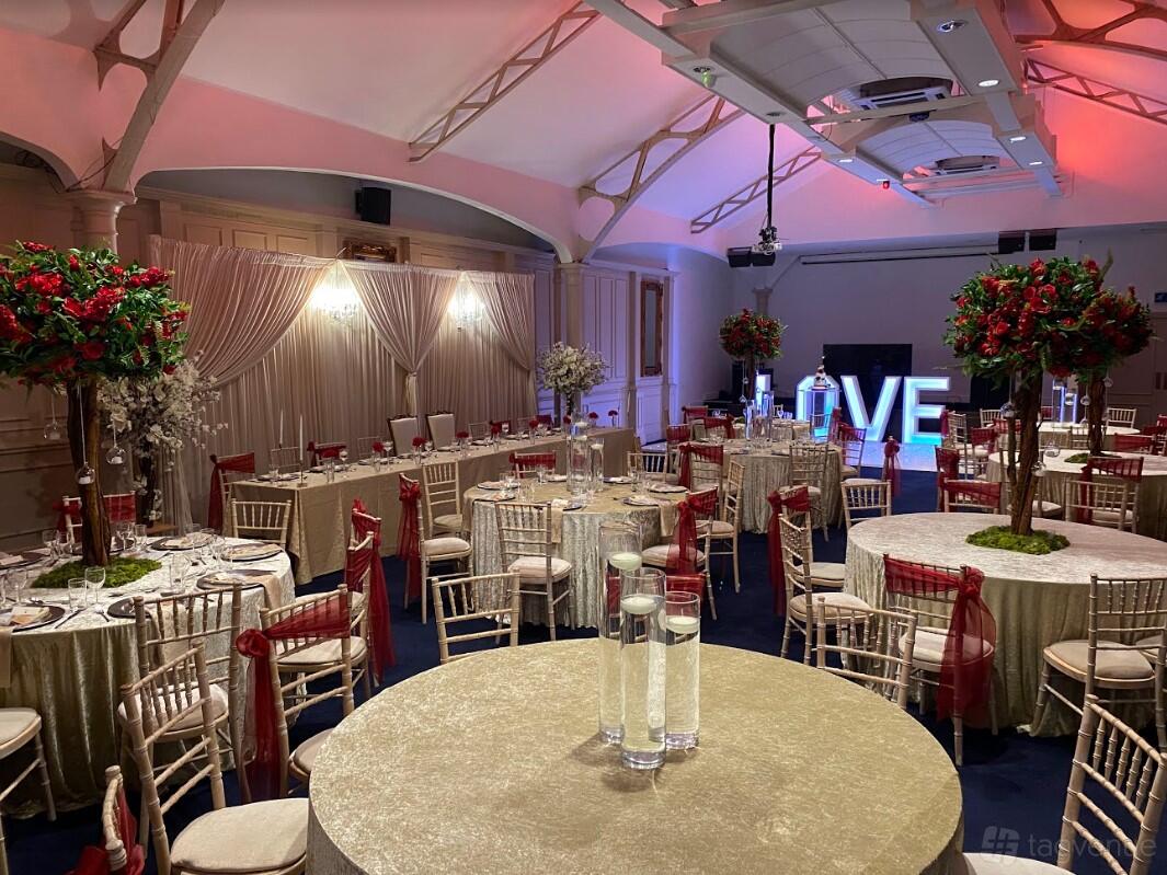A banquet hall with round tables, gold linens, red floral centerpieces, and illuminated LOVE letters at The Grand Station.