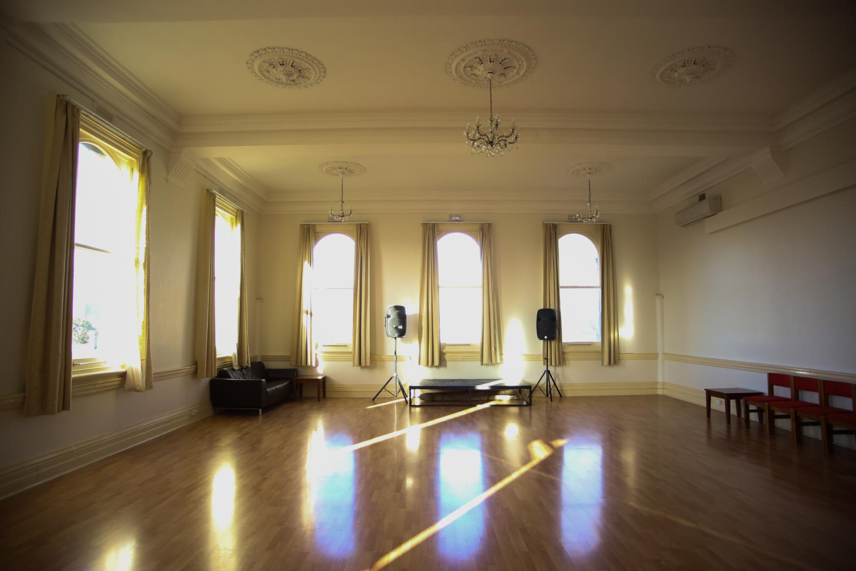 A function room with tall arched windows, chandeliers, and a small stage at The Russian House Emerald Hall.