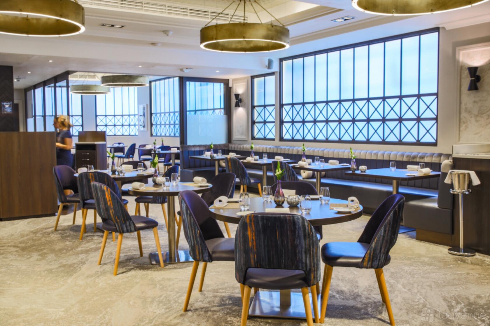 A restaurant with large industrial-style windows, round tables set with glassware, and cushioned booth seating at Adam's Restaurant.