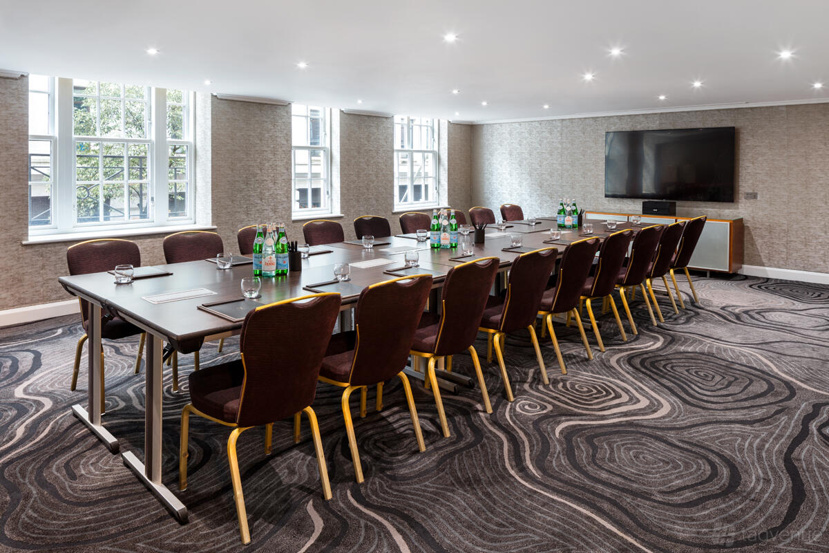 A meeting room with a boardroom table, upholstered chairs, large windows, and a wall-mounted screen at The May Fair Hotel.