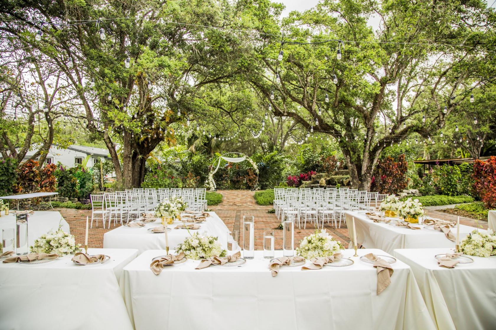 An outdoor event space with brick pathways, large shade trees, and white banquet tables at The Gardens of Weber Manor.