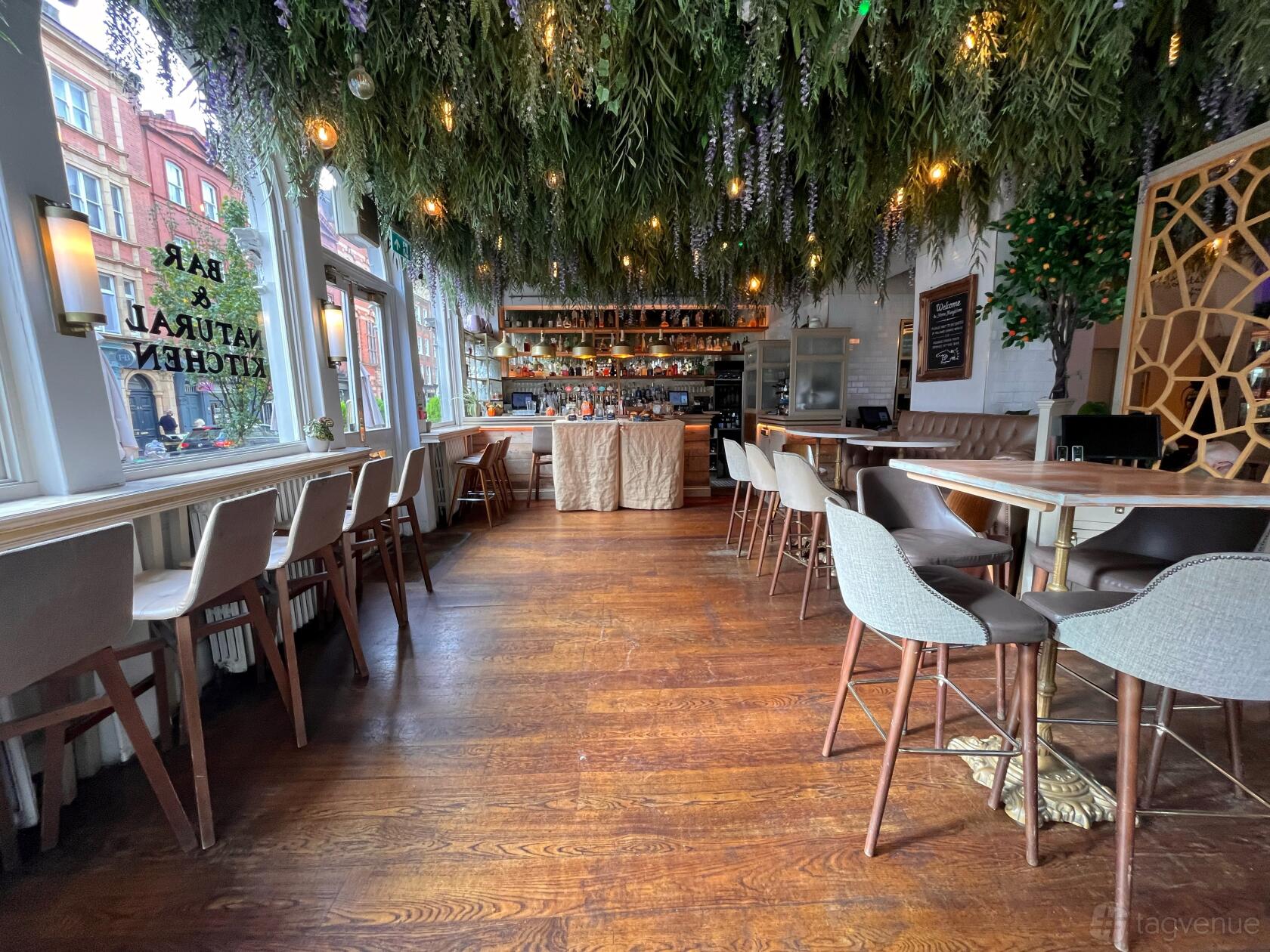 A bar in a restaurant with hanging greenery, wood floors, and high chairs around tables at Home Marylebone.