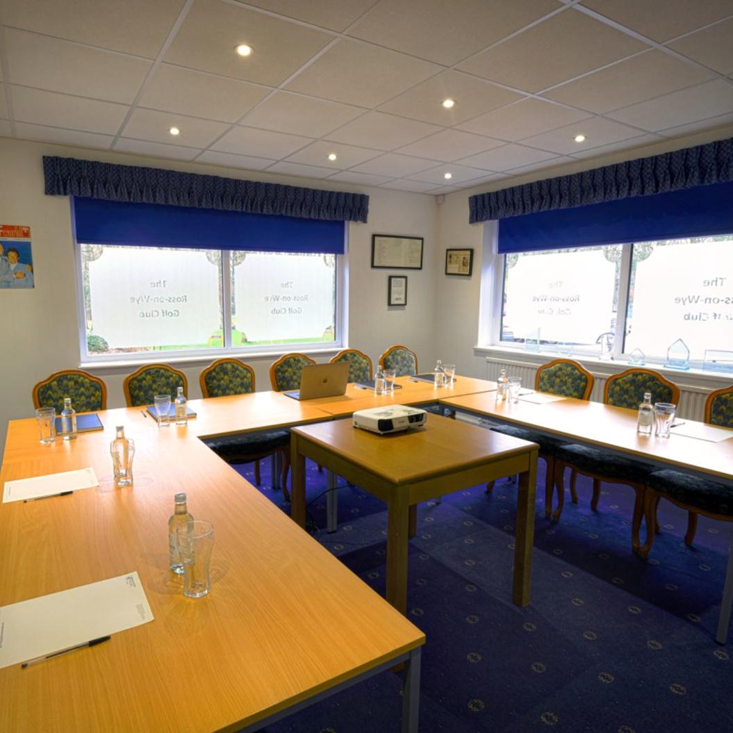 A meeting room with U-shaped wood tables, floral upholstered chairs, and large windows at The Ross-on-Wye Golf Club.