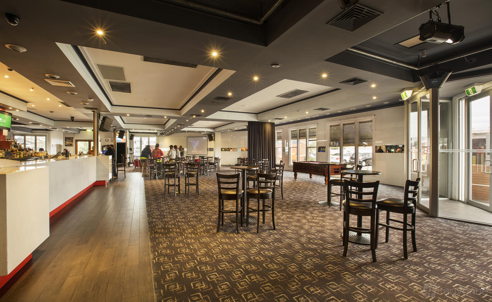 A function room in a pub with patterned carpet, scattered wooden chairs and tables, and large windows at Commercial Hotel South Morang.