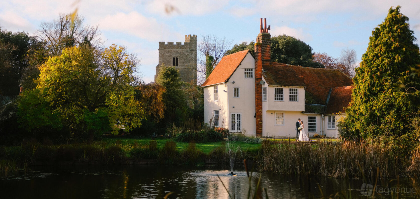 A historic hall with white walls, a red tile roof, and a pond with a fountain at East Mersea Hall.