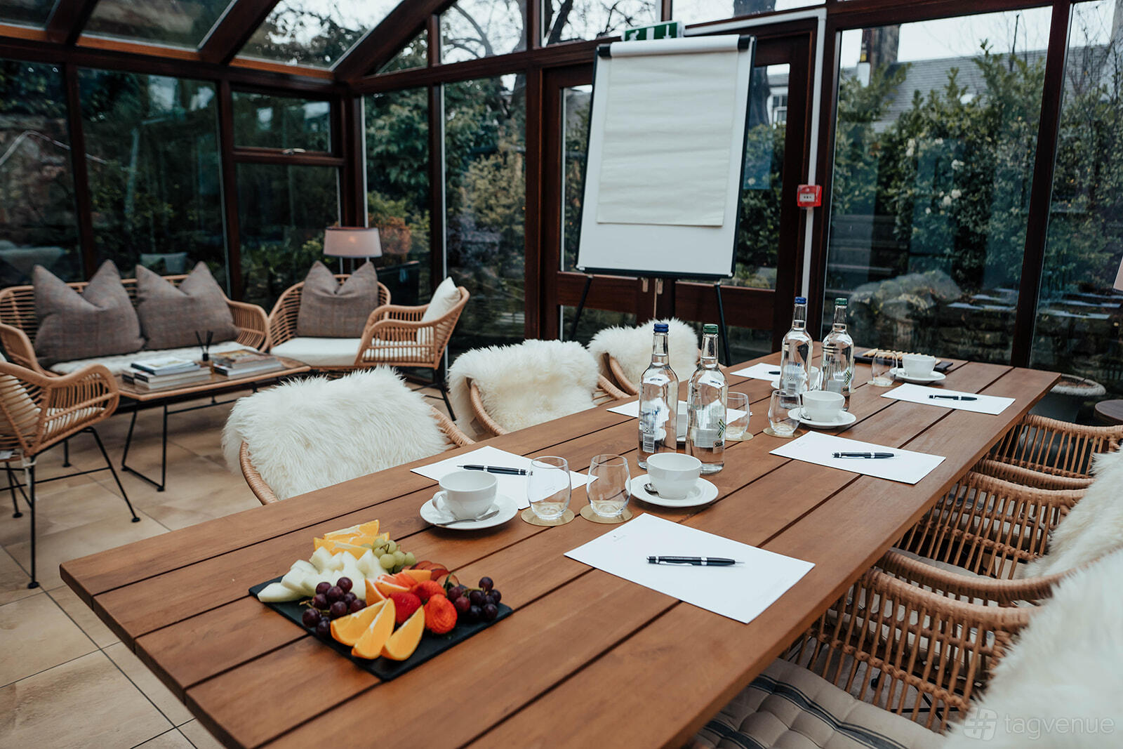 A conservatory in a hotel with glass walls, a wooden meeting table, and cushioned wicker chairs at The Roseate Edinburgh.