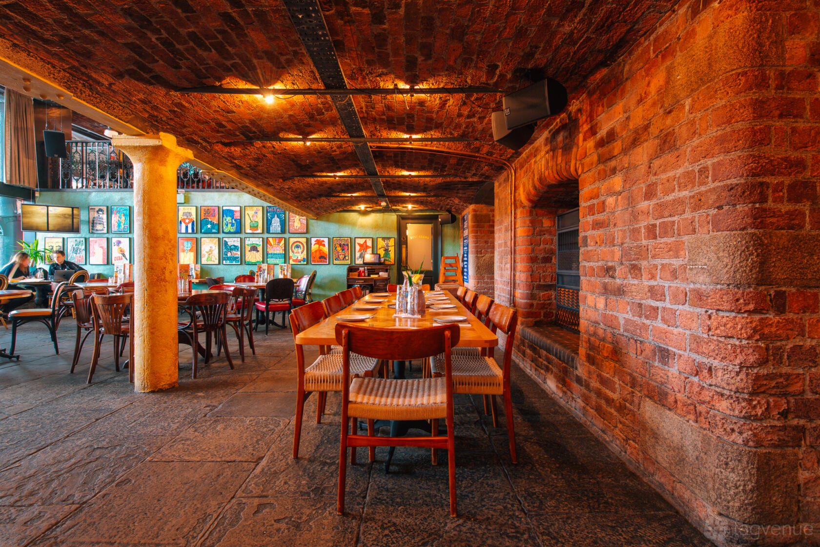 A bar with exposed brick walls, arched ceilings, wooden tables, and colorful framed artwork at Revolucion de Cuba Liverpool.