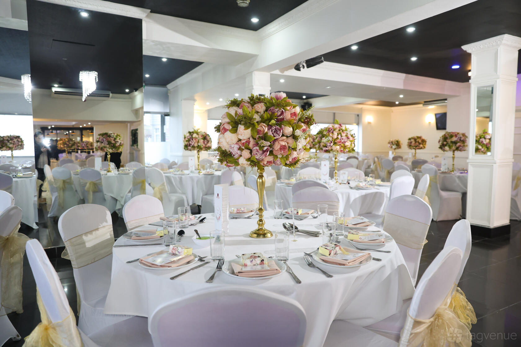 A banquet hall with round tables draped in white linens, gold chair sashes, and tall floral centerpieces at Crown Banqueting Suite (Ilford).
