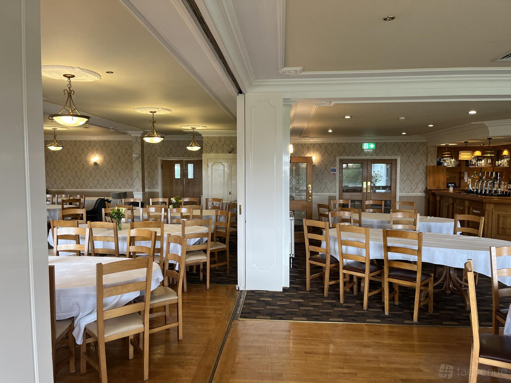 A function room with round tables covered in white linens, wooden chairs, and a bar area at The Manor Golf Club.
