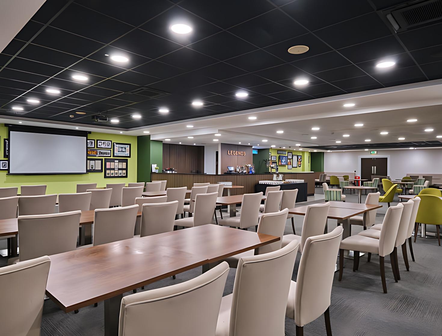 A stadium lounge with rows of tables and cream chairs, a large projector screen, and framed sports memorabilia at Swansea.com Stadium.