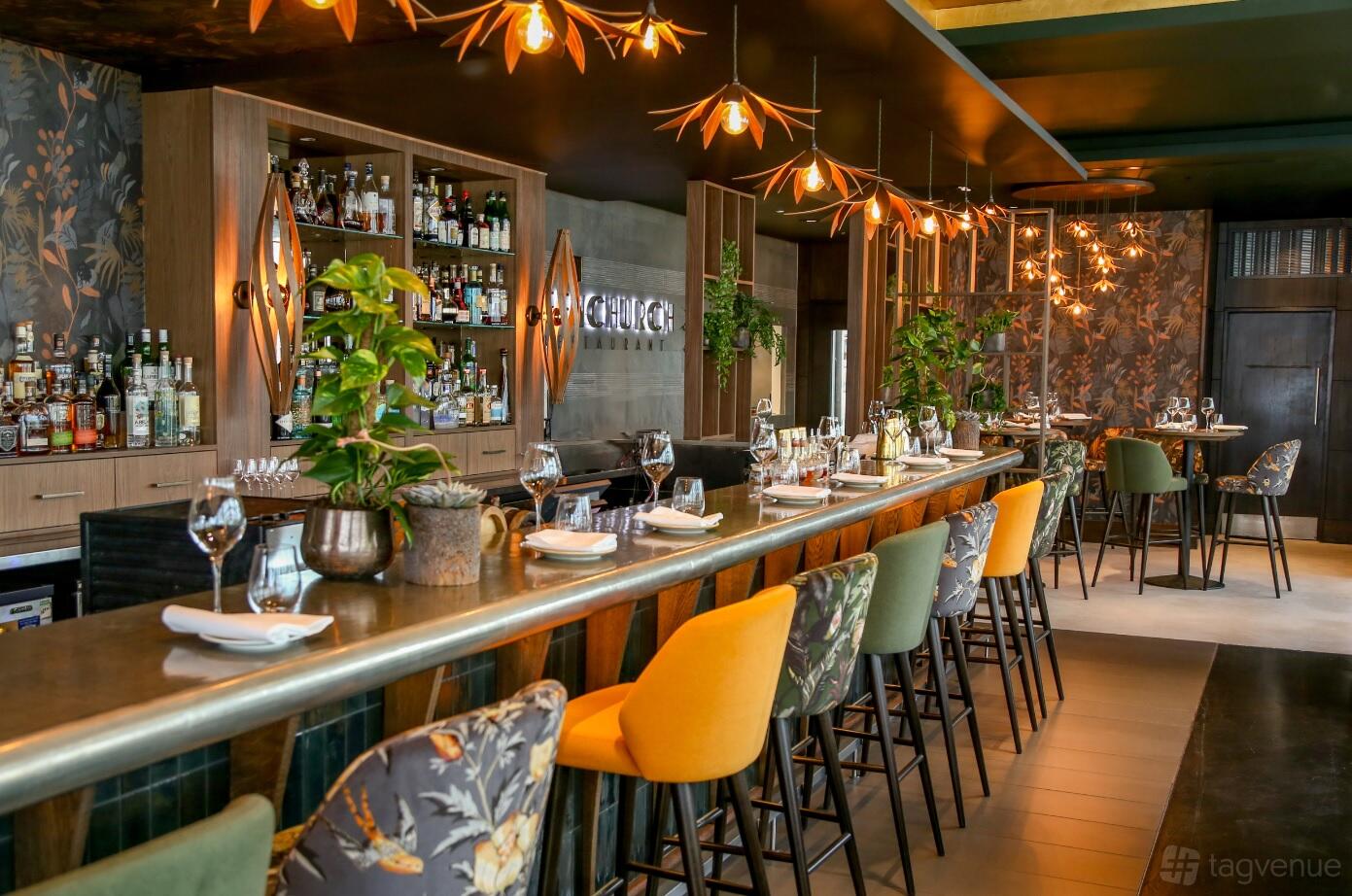 An event venue with a long bar, floral upholstered chairs, pendant lights, and lush potted plants at Sky Garden.