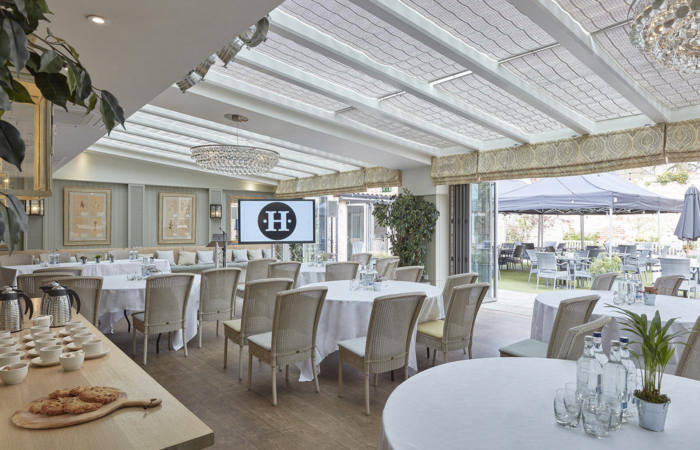 An event space with glass ceiling, chandeliers, round tables, and garden views at The Hoste Arms.