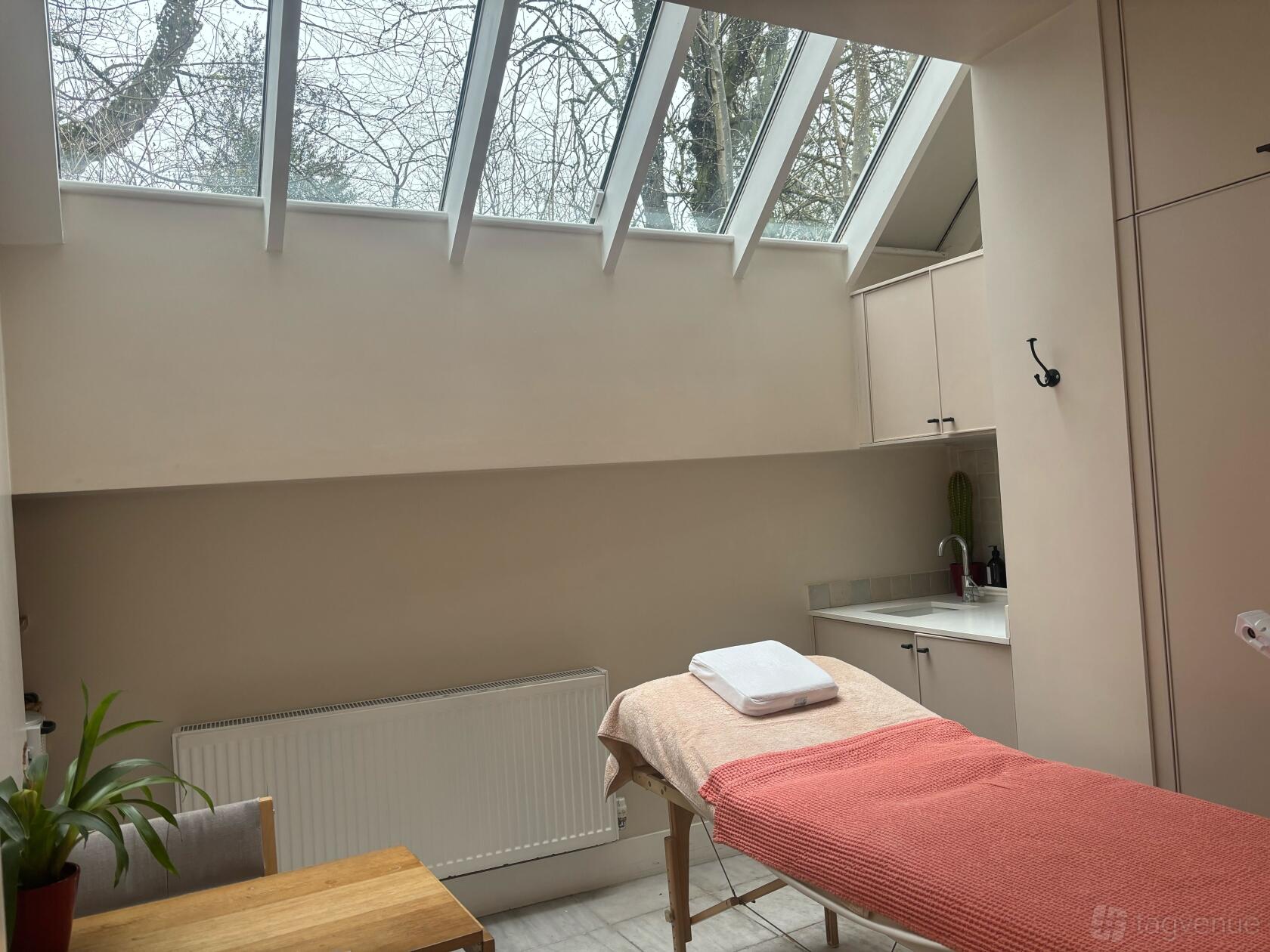 A therapy room with a skylight, treatment bed with a coral blanket, and built-in cabinets at The Hearth.