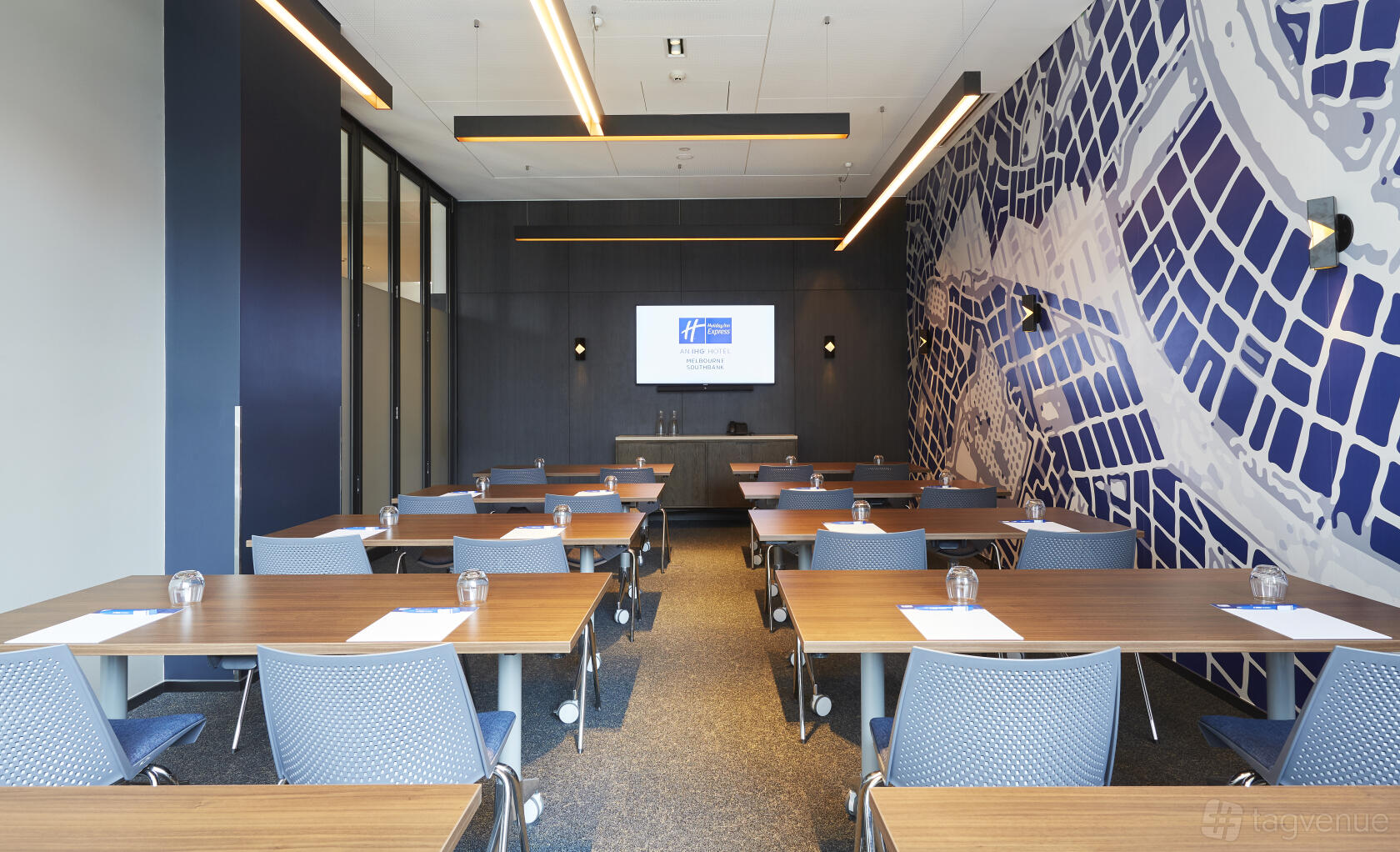 A meeting room with wood tables, blue chairs, large wall mural, and a presentation screen at Holiday Inn Express Melbourne Southbank.