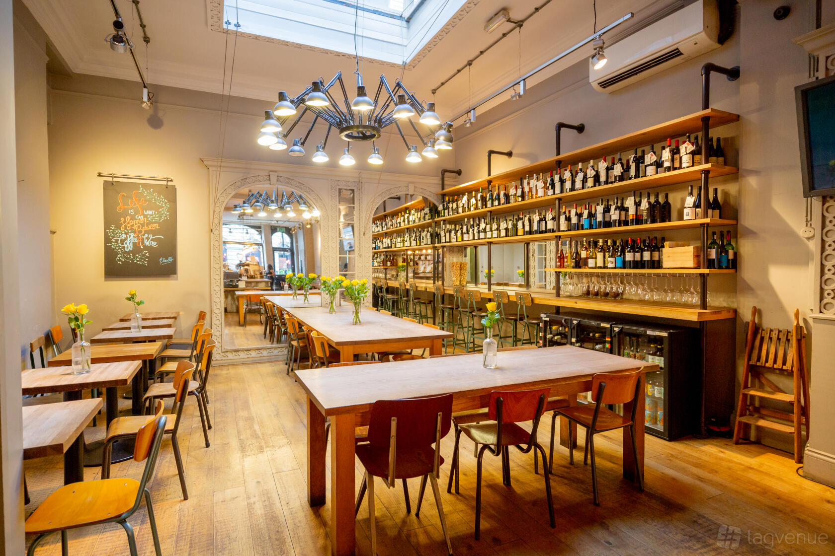 A cafe with wooden tables, chairs, flower centerpieces, and a wall of shelves stocked with bottles at Notes Coffee & Bars Trafalgar square.