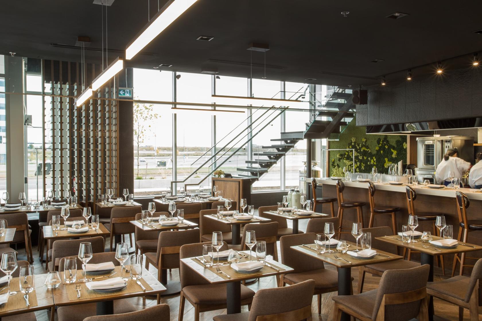 A restaurant with floor-to-ceiling windows, set tables with glassware, and an open kitchen at Terre Rouge Craft Kitchen.