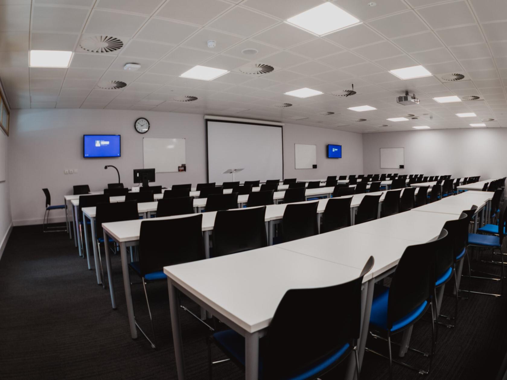 A meeting room with rows of white tables, black chairs, large projector screens, and whiteboards at University of Law - Leeds.