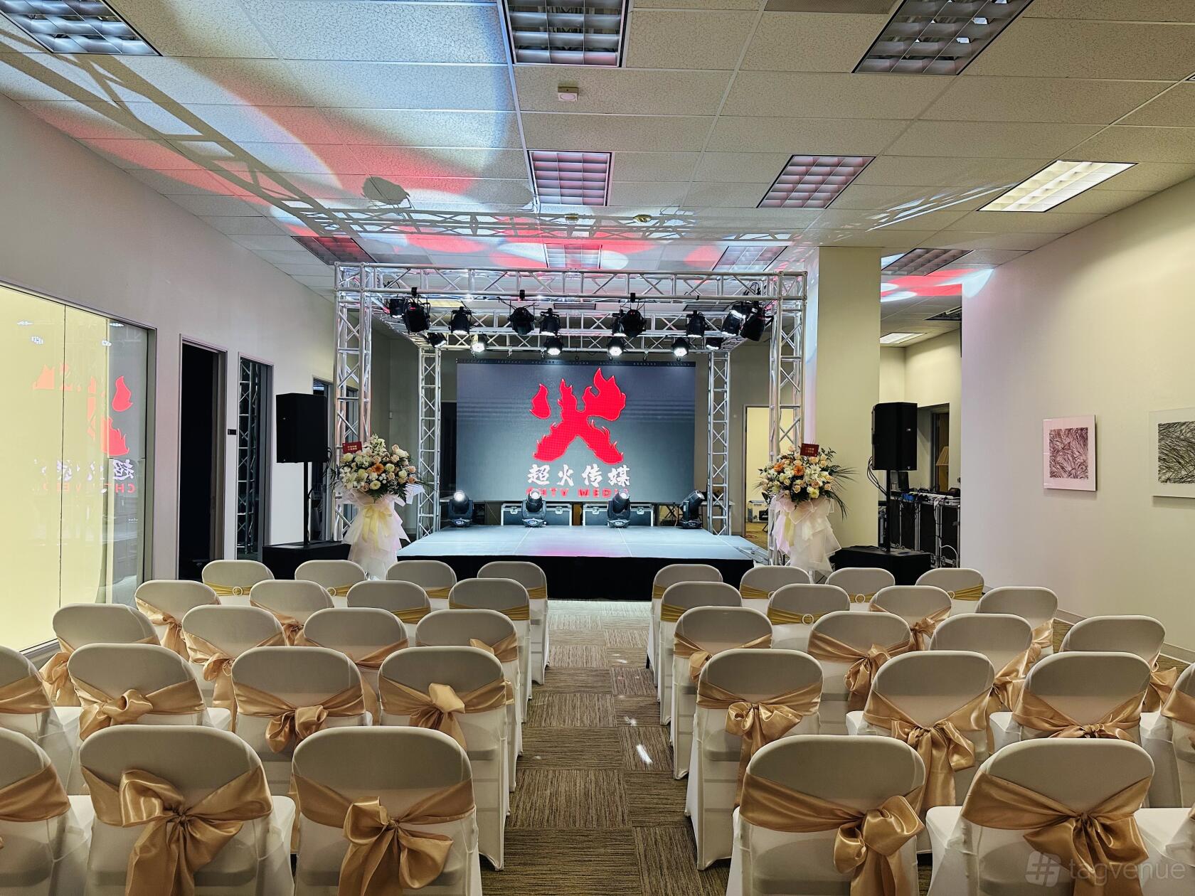An event space with rows of white chairs tied with gold ribbons facing a stage with lighting truss at CHTV Media & Entertainment LLC.