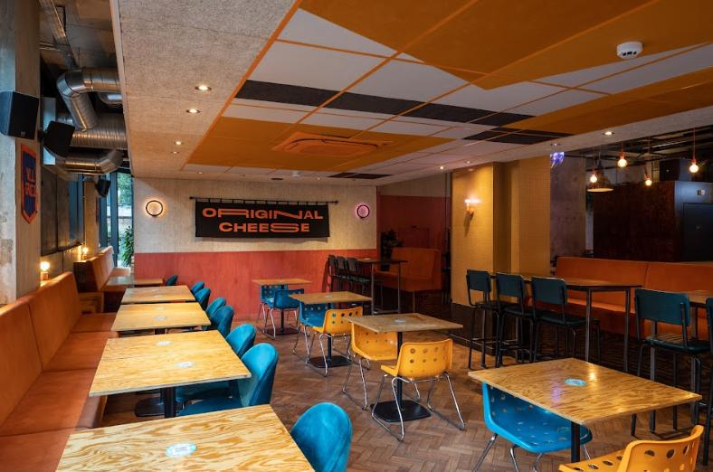 An event space in a restaurant with wooden tables, colorful chairs, orange banquette seating, and exposed ductwork at Nell's at Kampus.