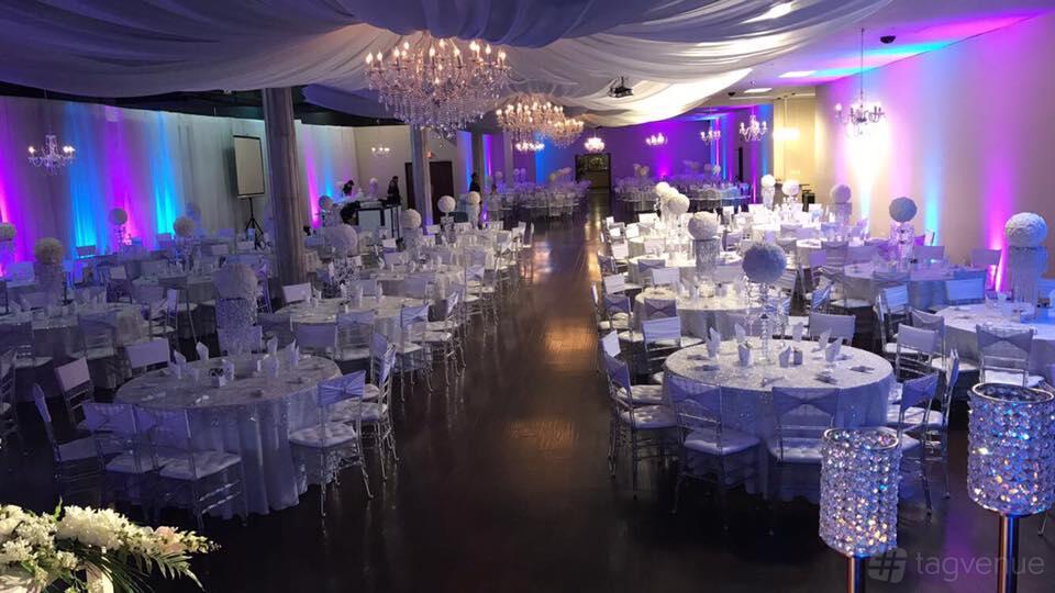 A banquet hall with draped ceilings, crystal chandeliers, uplighting, and round tables set with white linens at Las Lomas Irving Banquet Hall.