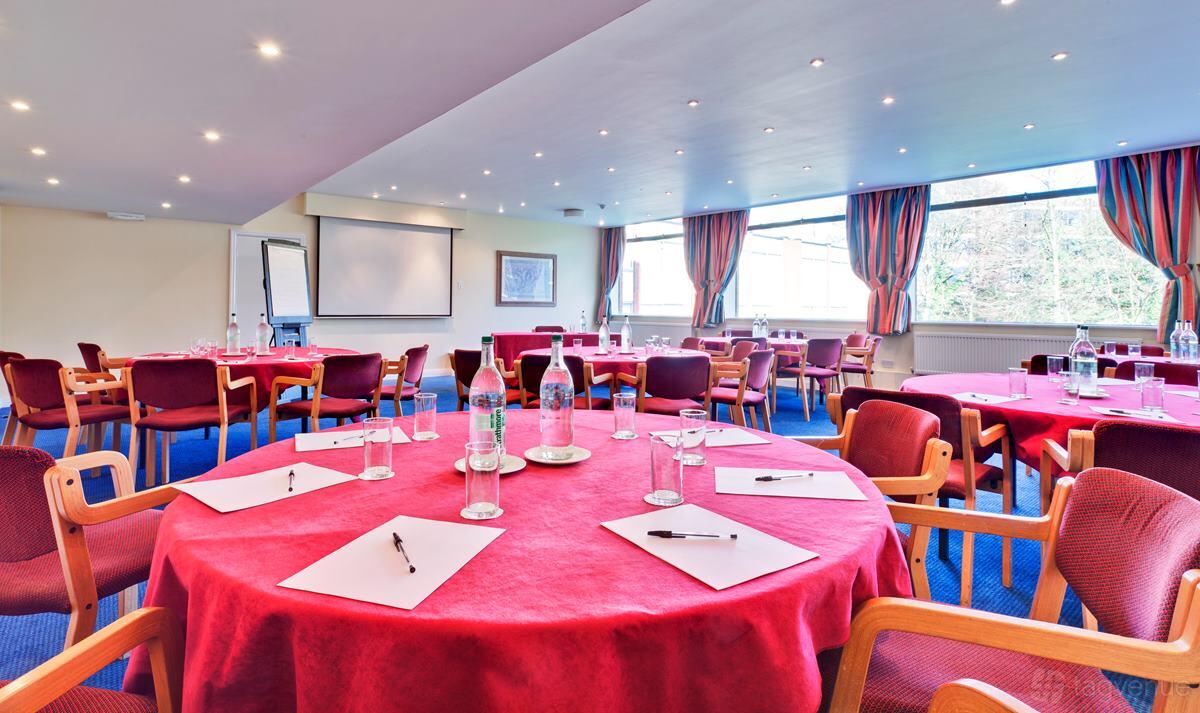 A hotel function room with round tables covered in red linens, notepads, water bottles, and large windows at Plough & Harrow Hotel.