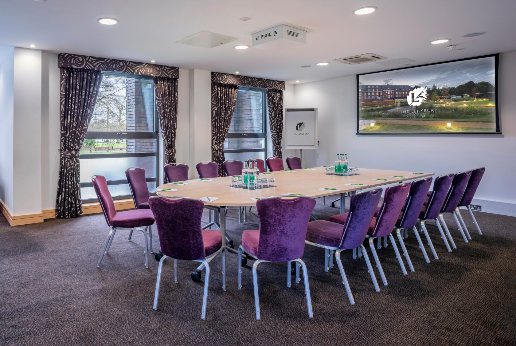 A meeting room with a U-shaped table, purple chairs, large windows, and a wall-mounted screen at The Lensbury.