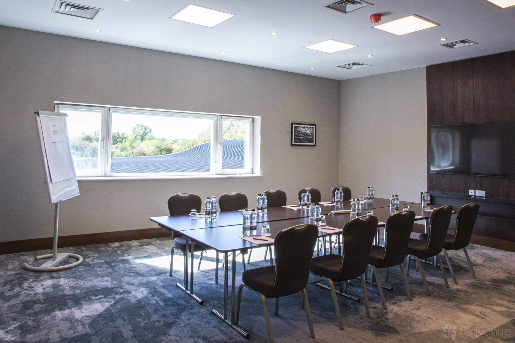 A hotel meeting room with a rectangular table, bottled water, notepads, a large window, and a flip chart at Crowne Plaza Marlow.
