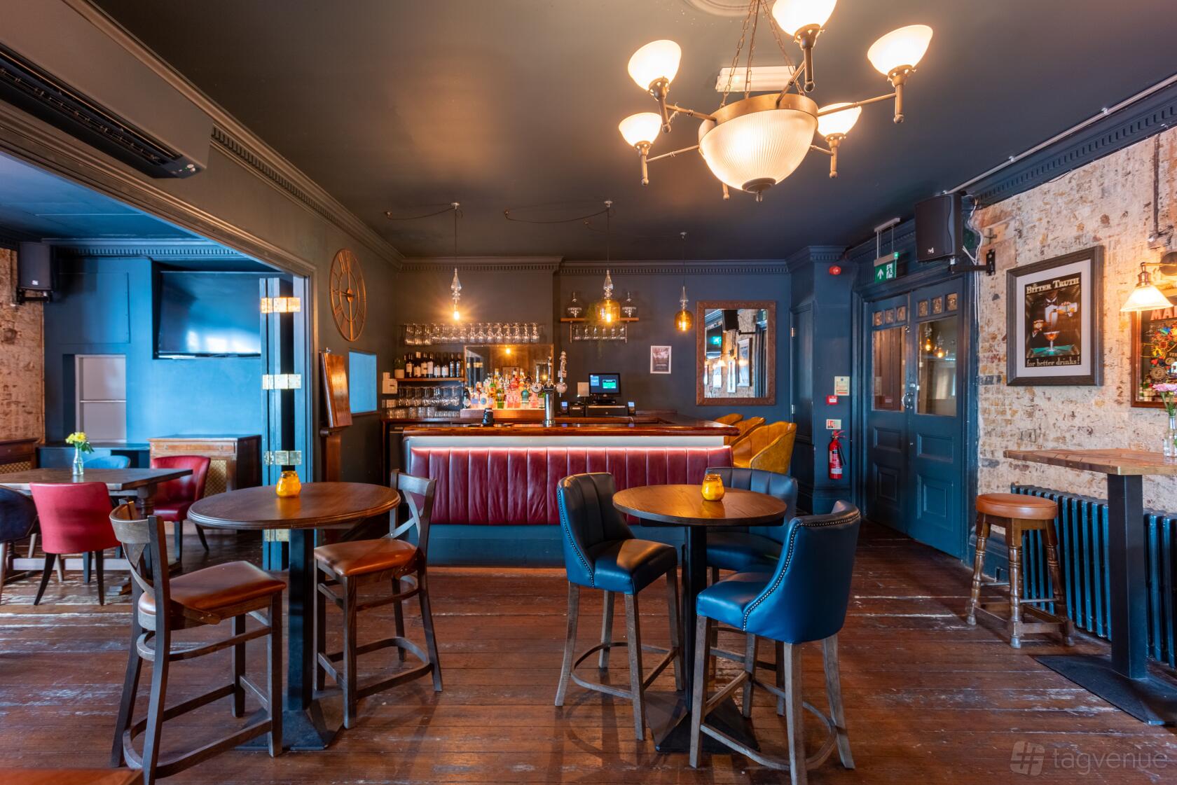 An event space in a pub with exposed brick walls, round wooden tables, and cushioned chairs at The Wheatsheaf Tooting.