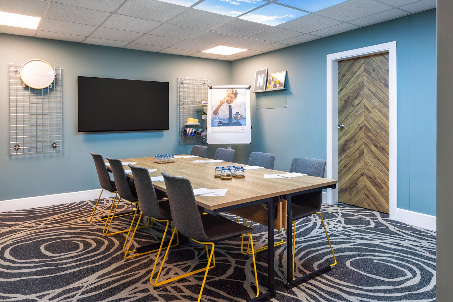 A meeting room with a rectangular table, gray chairs, wall-mounted screen, and modern patterned carpet at Novotel Edinburgh Centre.