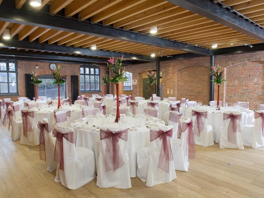 An event venue with exposed brick walls, wood beam ceiling, and round tables with white linens and purple chair sashes at IET Birmingham: Austin Court.