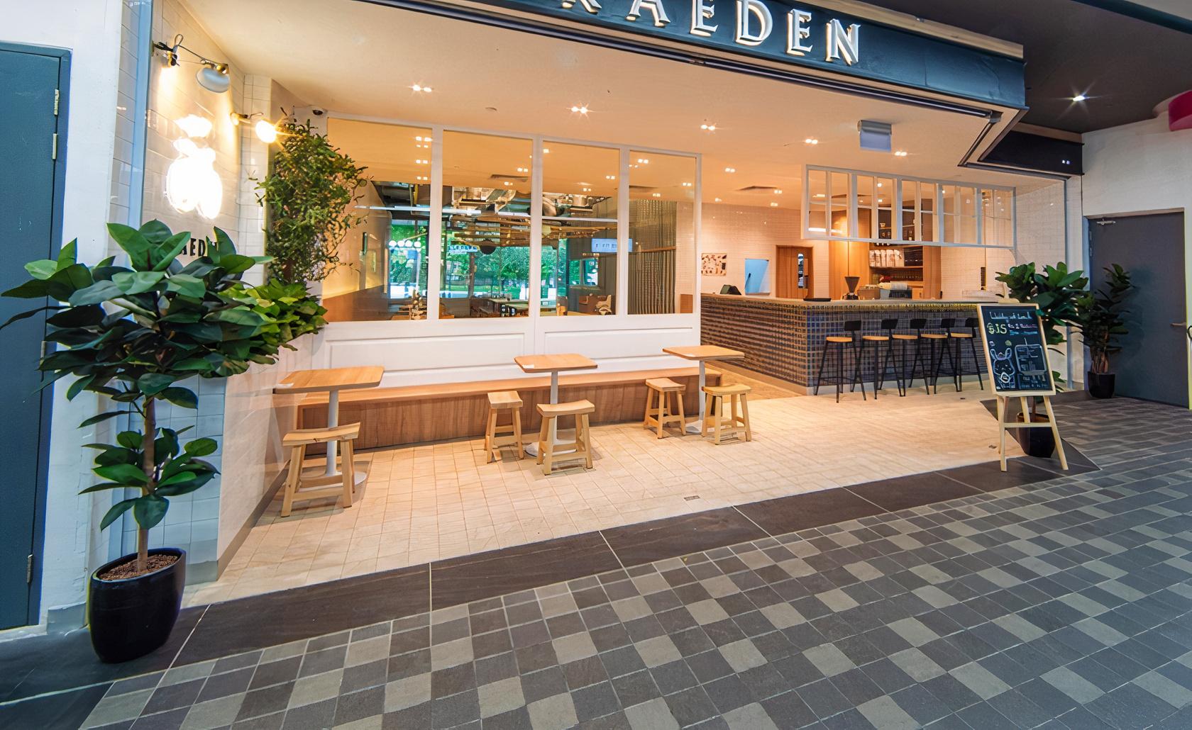 A café with light wood tables, a white bench, and potted plants at the entrance of Kaeden Cafe.