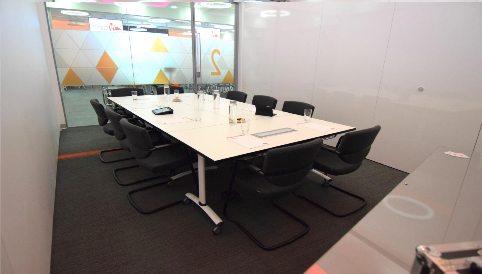 A meeting room with a long white conference table, black chairs, and glass walls with geometric patterns at Millennium Point.