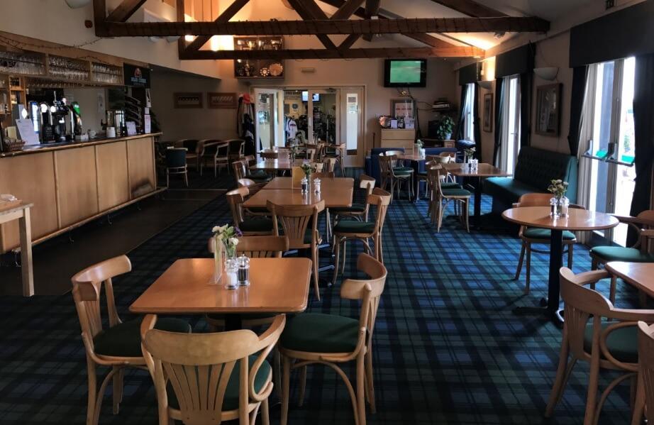 An event venue with wooden beams, tartan carpet, and wooden tables with small floral centerpieces at Rivenhall Oaks Golf Centre.