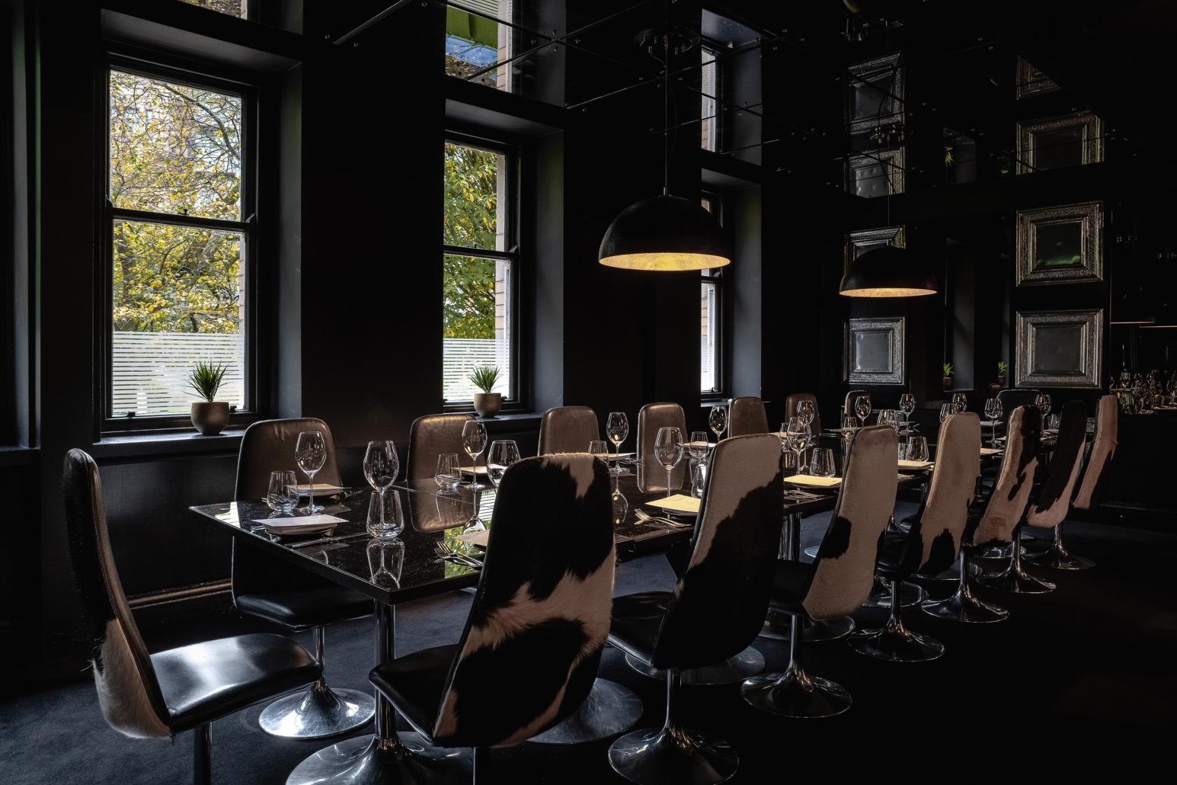 A dining room with cowhide chairs, dark decor, and set tables by large windows at Gaucho Manchester.