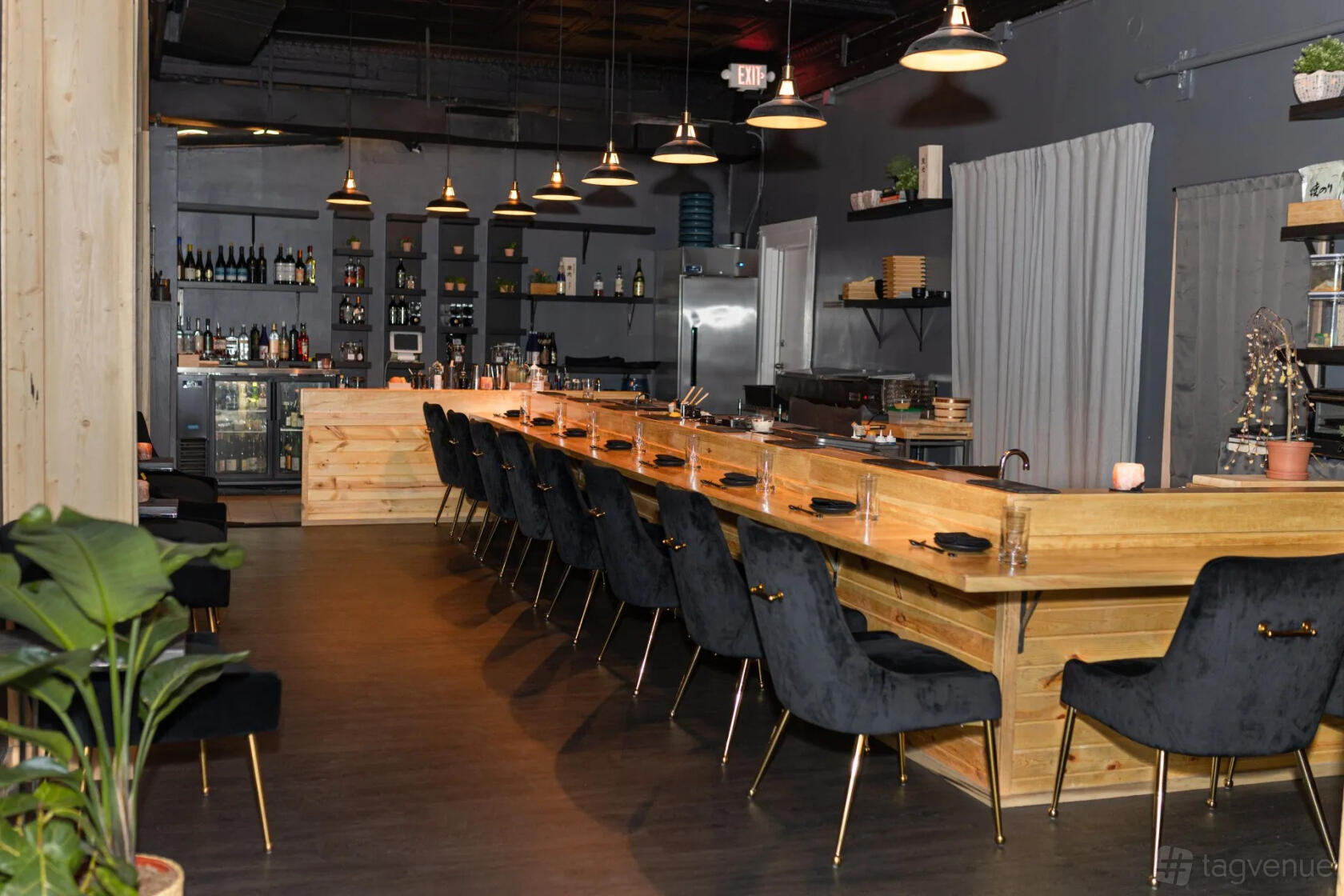 A restaurant with a long wooden dining counter, black upholstered chairs, and open shelving at AJI.