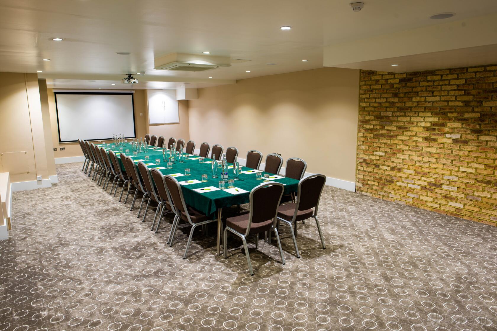 An event space with U-shaped tables, green tablecloths, and a yellow brick accent wall at The Wesley Euston Hotel.