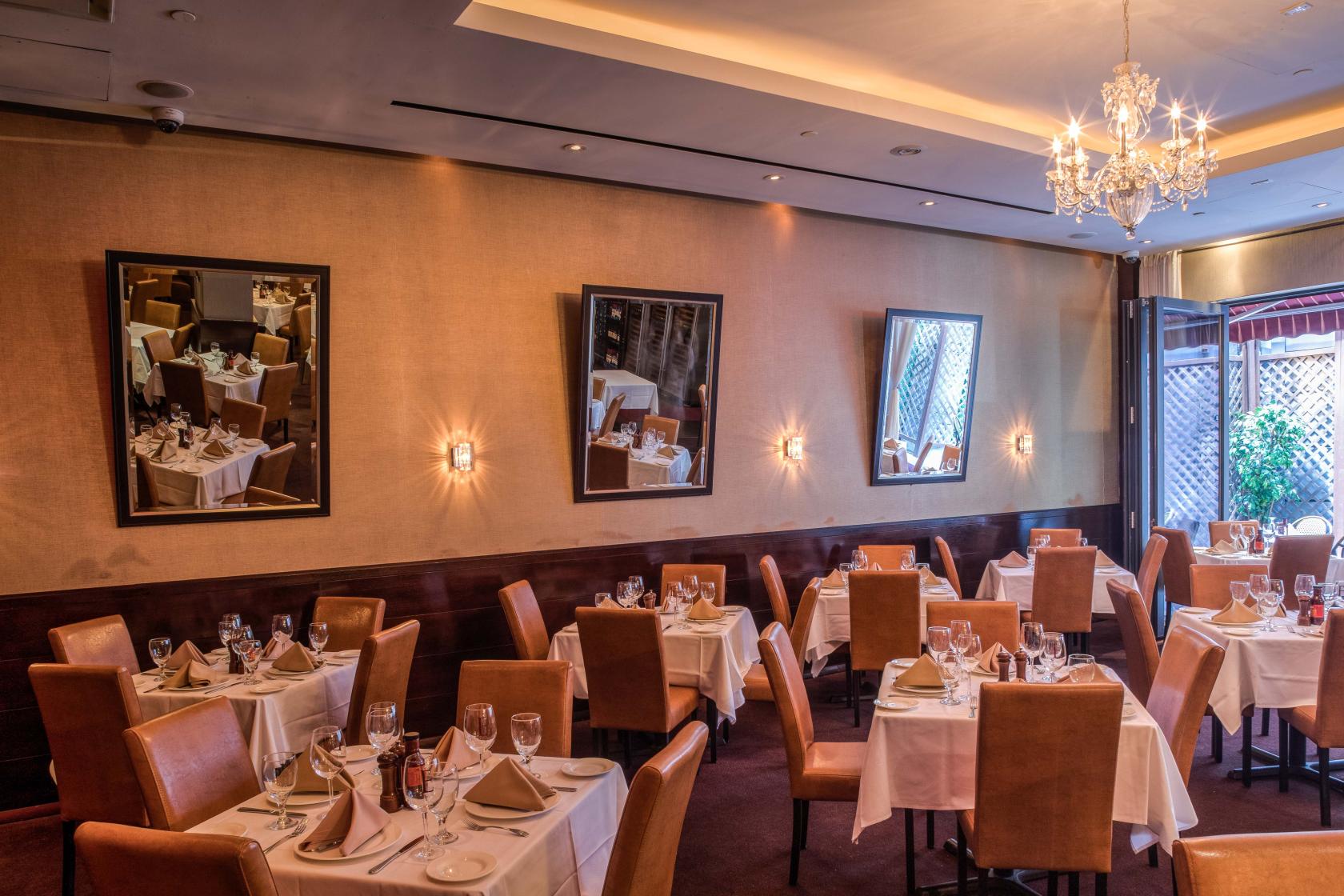 A restaurant dining room with tan leather chairs, white tablecloths, wall sconces, and a crystal chandelier at Empire Steak House West.