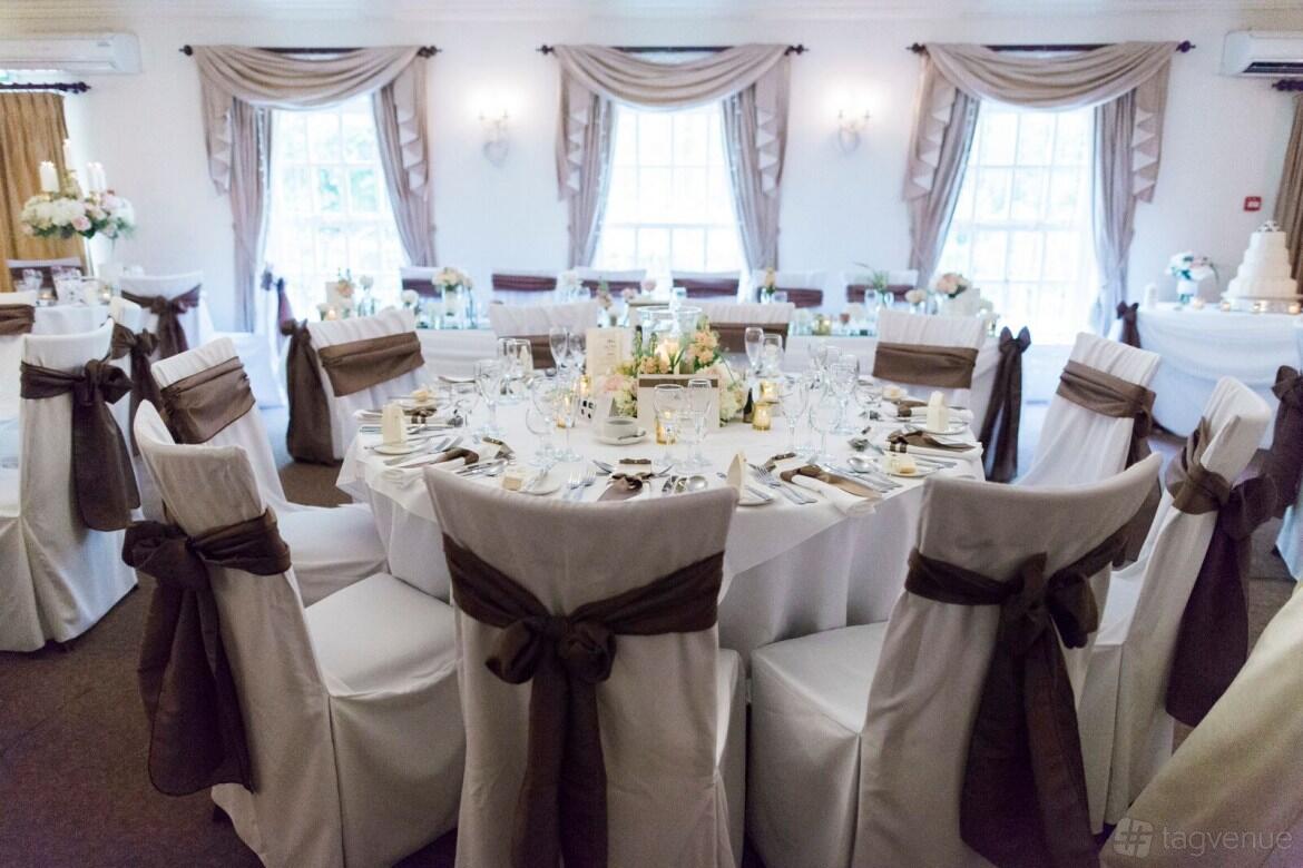A country house with round tables set for dining, white chairs with brown sashes, and large windows at Singleton Lodge Country House Hotel.