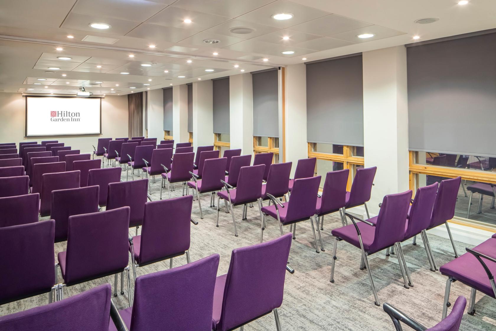 A meeting room with rows of purple chairs, carpeted floors, and ceiling spotlights at Hilton Garden Inn Birmingham.
