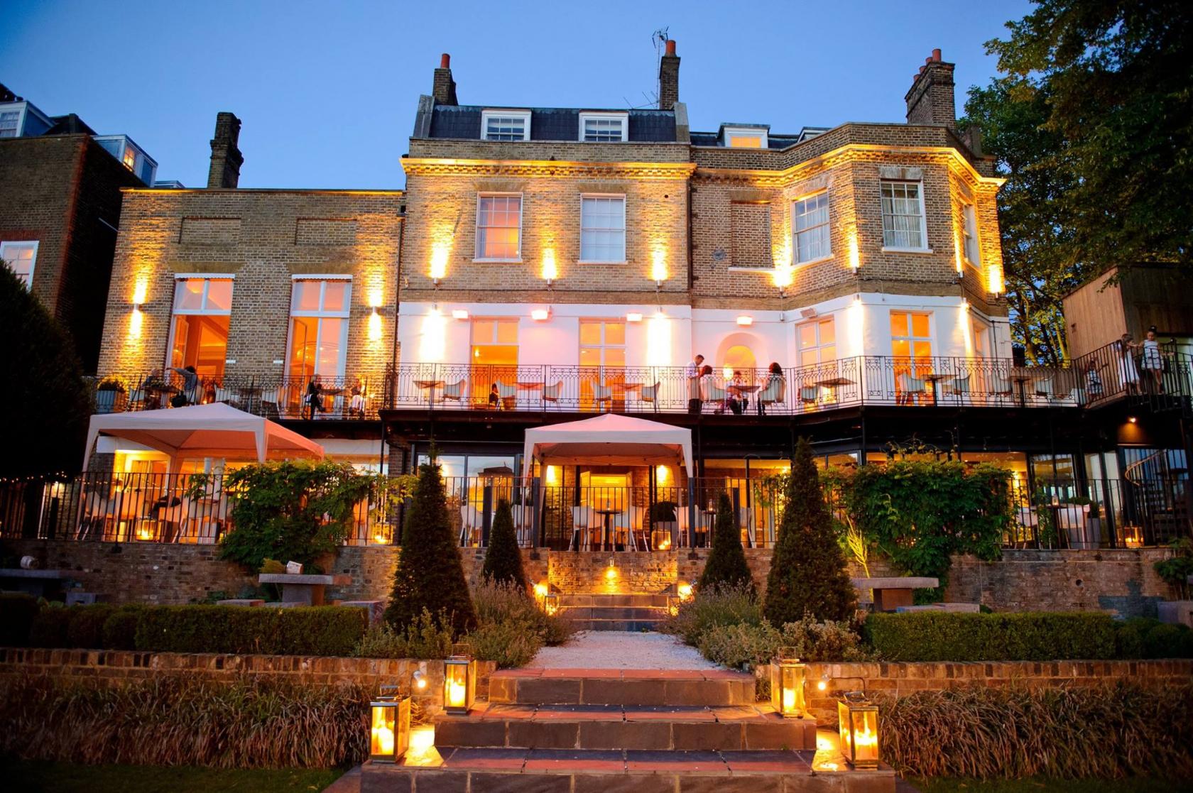 A hotel with an illuminated brick facade, large windows, and a terrace overlooking a landscaped garden at Bingham Riverhouse.
