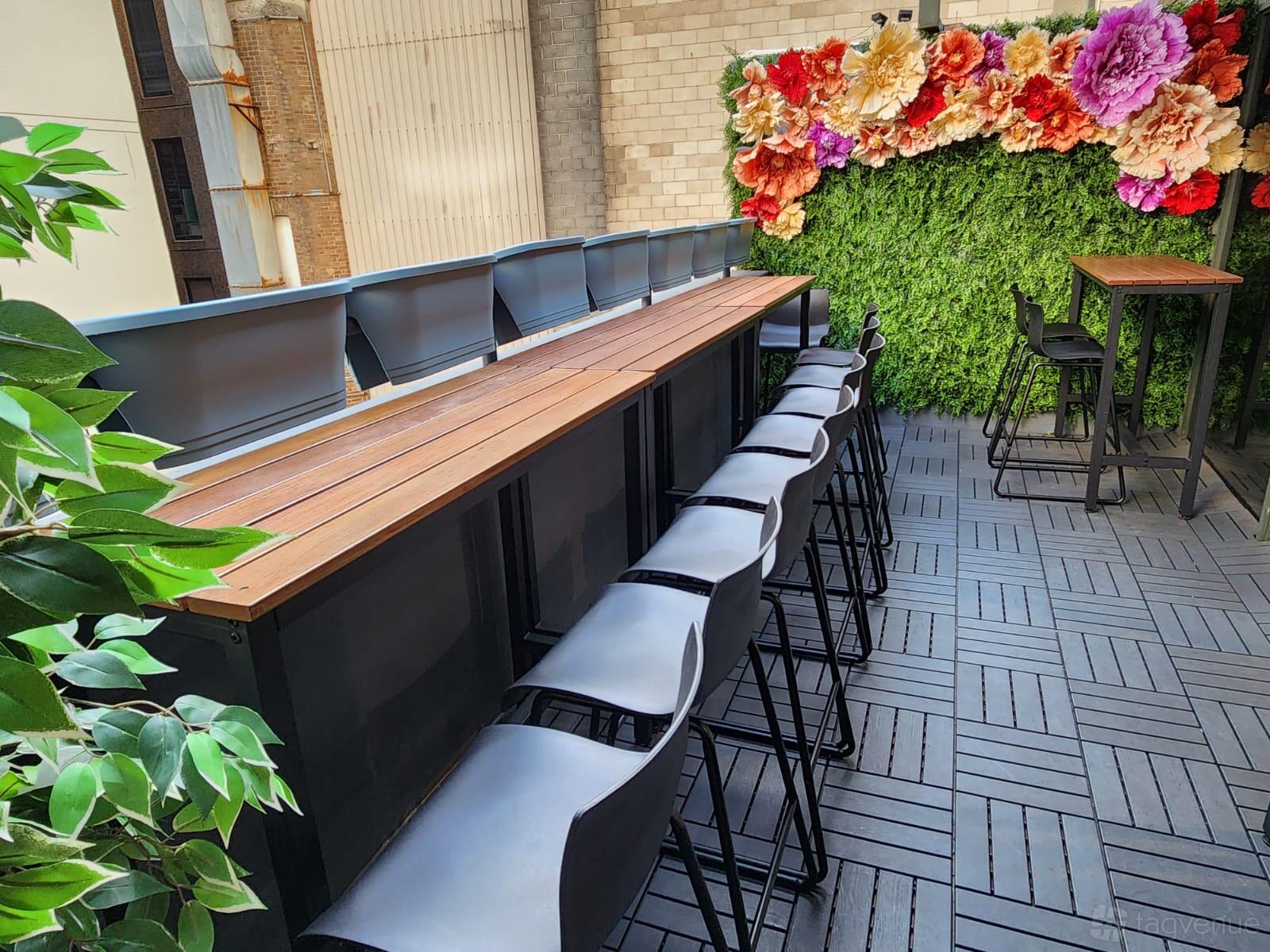 A loft event space with a long wooden bar, black stools, and a flower wall on the large balcony at Loft on George.
