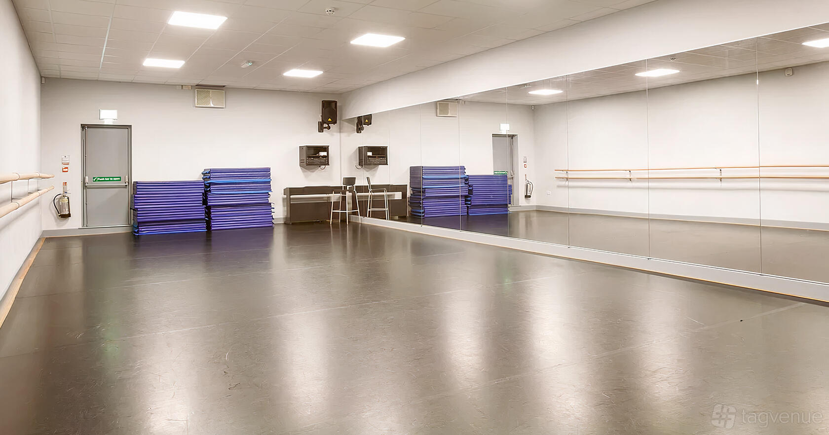 A dance studio with a wall of mirrors, ballet barres, and stacked exercise mats at Adagio School Of Dance.