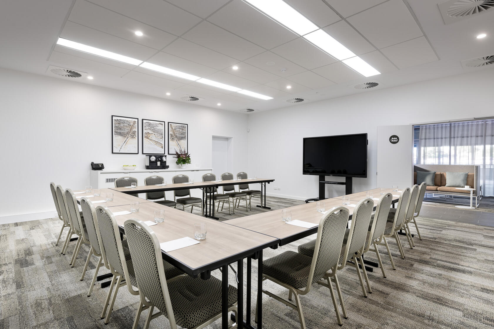 A hotel meeting room with U-shaped tables, cushioned chairs, wall art, and a large screen at Quest Midland.