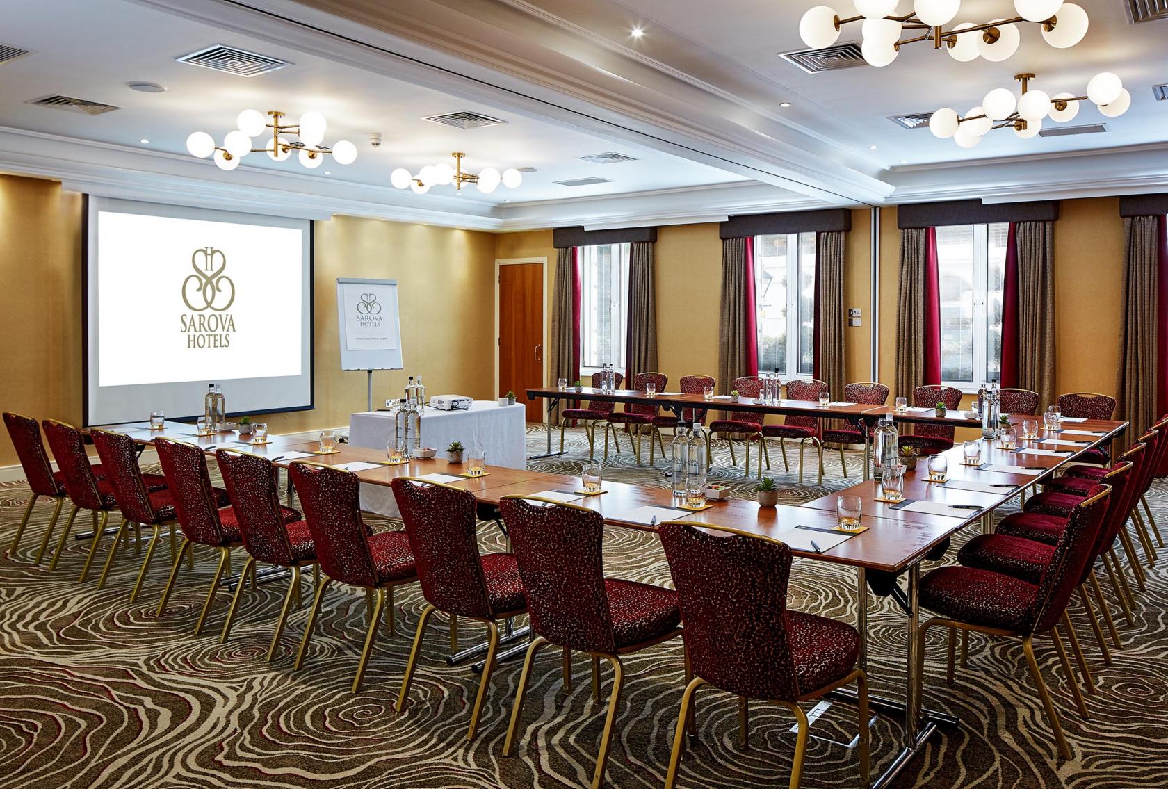 A meeting room with U-shaped tables, red upholstered chairs, and large windows at The Rembrandt Hotel.