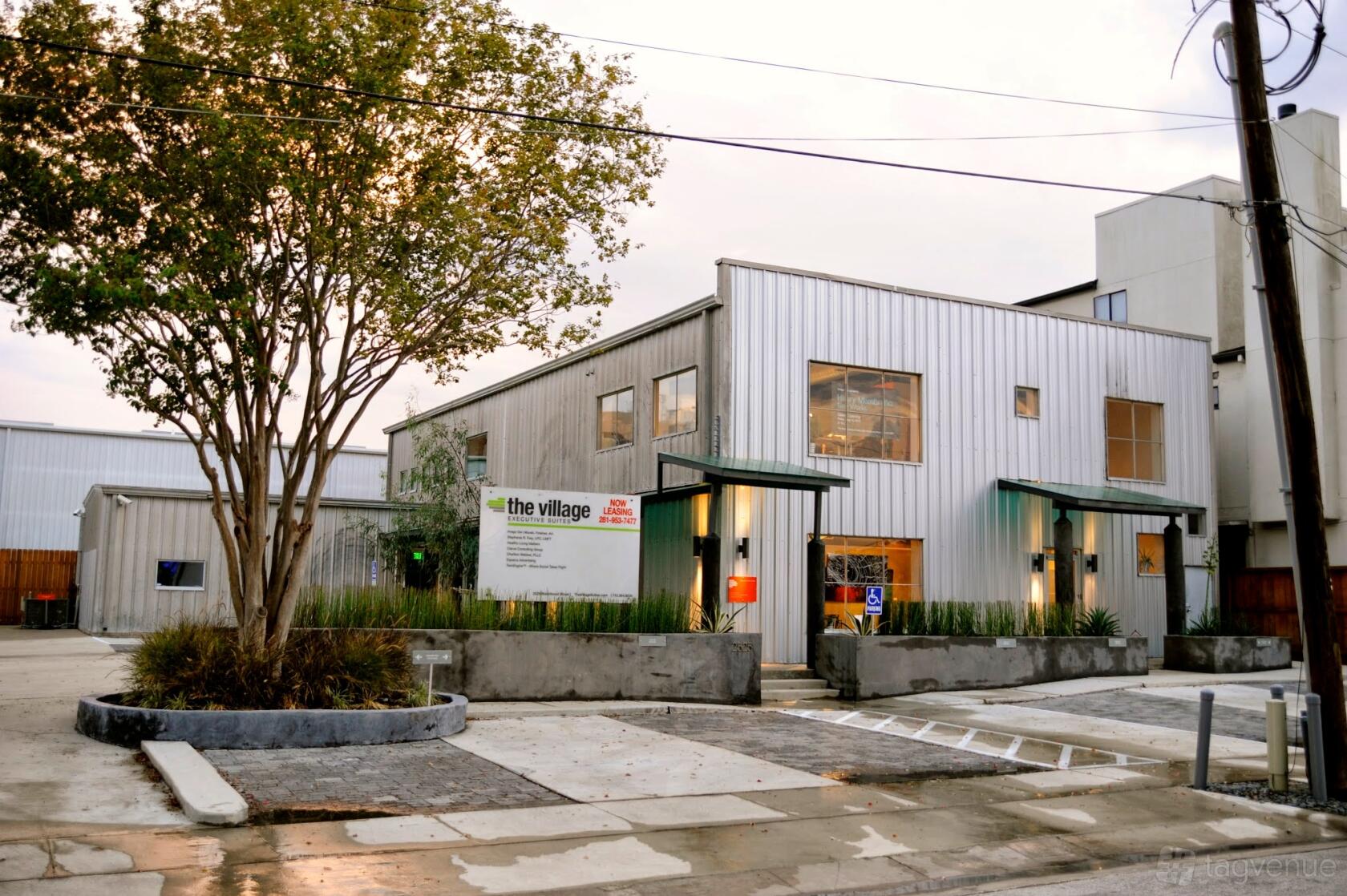 A coworking space with a metal facade, large windows, and a landscaped entrance at The Village Suites.