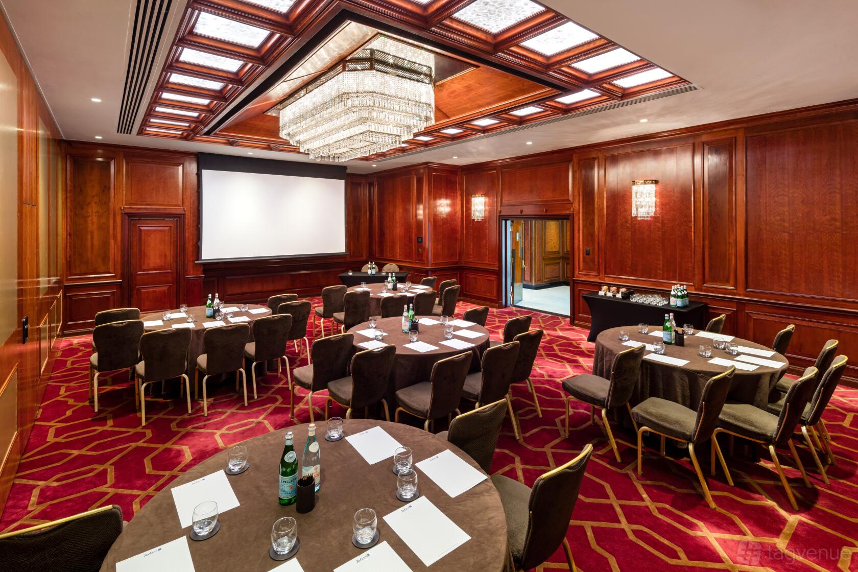 An auditorium with wood-paneled walls, a crystal chandelier, and round tables set for a meeting at Radisson Blu Hotel, London Heathrow.