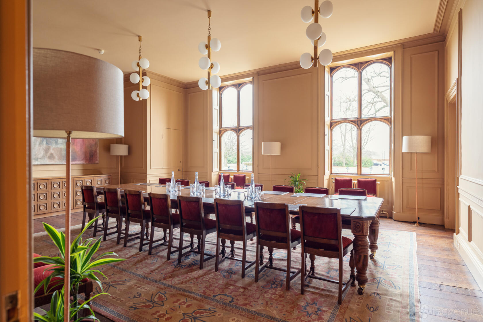 A meeting room with arched windows, a long wooden table, glass water bottles, and vintage chairs at Ashridge House.